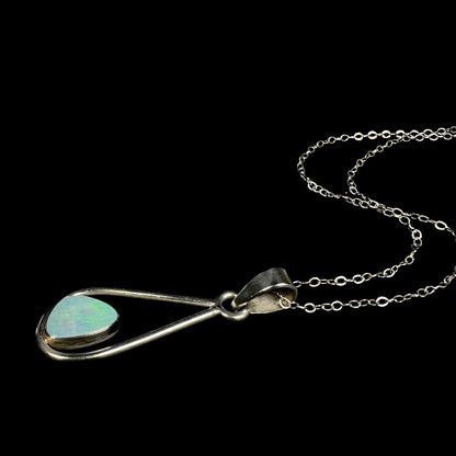 A sterling silver opal doublet necklace shown laying on a black background.  The opal has soft green play of color under cool studio light.