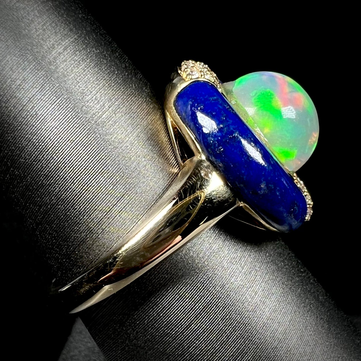 Side close-up view of a solid 14k yellow gold Ethiopian opal and lapis lazuli women's ring on a display.  The ring's shank is in clear focus.