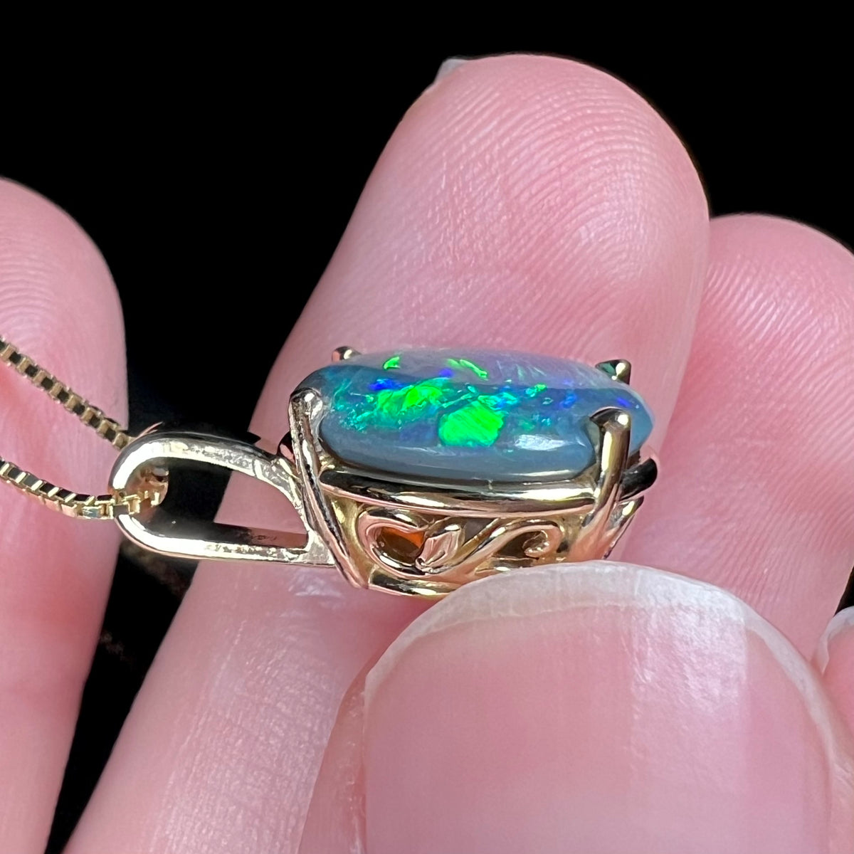 Side view of a solid Australian black opal pendant; the pendant has scrolling 14k gold vine and leaf filigree design.  The opal has green flashes and dark gray potch matrix