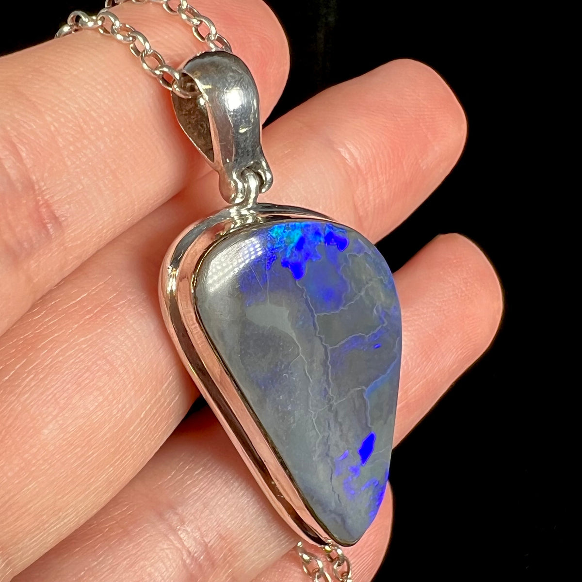 Angled macro view of solid Australian black opal pendant in sterling silver held in a hand, blue flashes seen in the opal