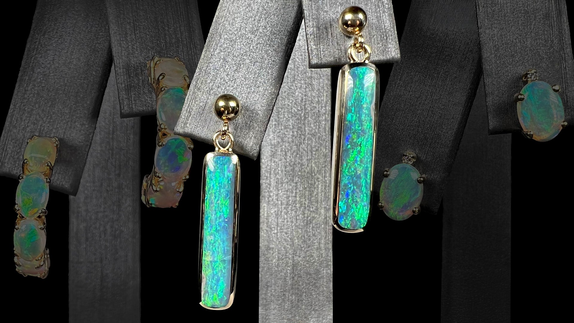 A pair of solid yellow gold Australian opal dangle earrings shown on an earring display in front of a dark backround showing faded opal semi-hoop earrings and opal stud earrings.