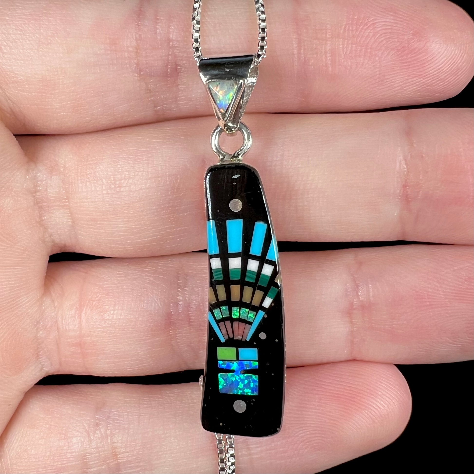 Close-up of Southwest-style opal and black onyx inlay pendant with kachina in headdress motif, held in hand under studio light.