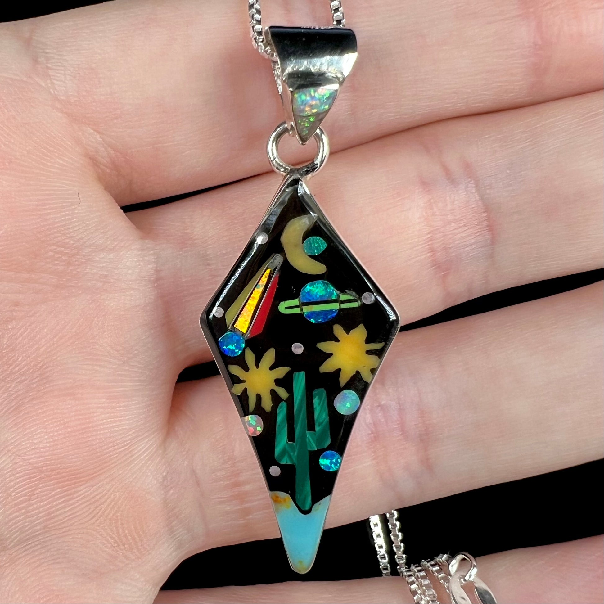 Close-up of Southwest-style opal inlay pendant with desert and space motif held in hand under cool studio light.