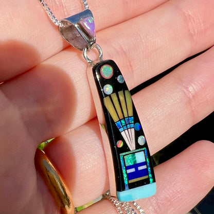Angled close-up of Southwest-style opal and black onyx inlay pendant held in hand under natural light.  The opals flash with different colors.