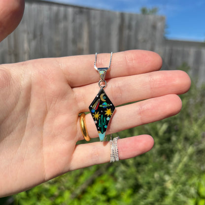 Hand holding sterling silver inlay necklace with outer-space design in natural light against blurred outdoor backdrop.