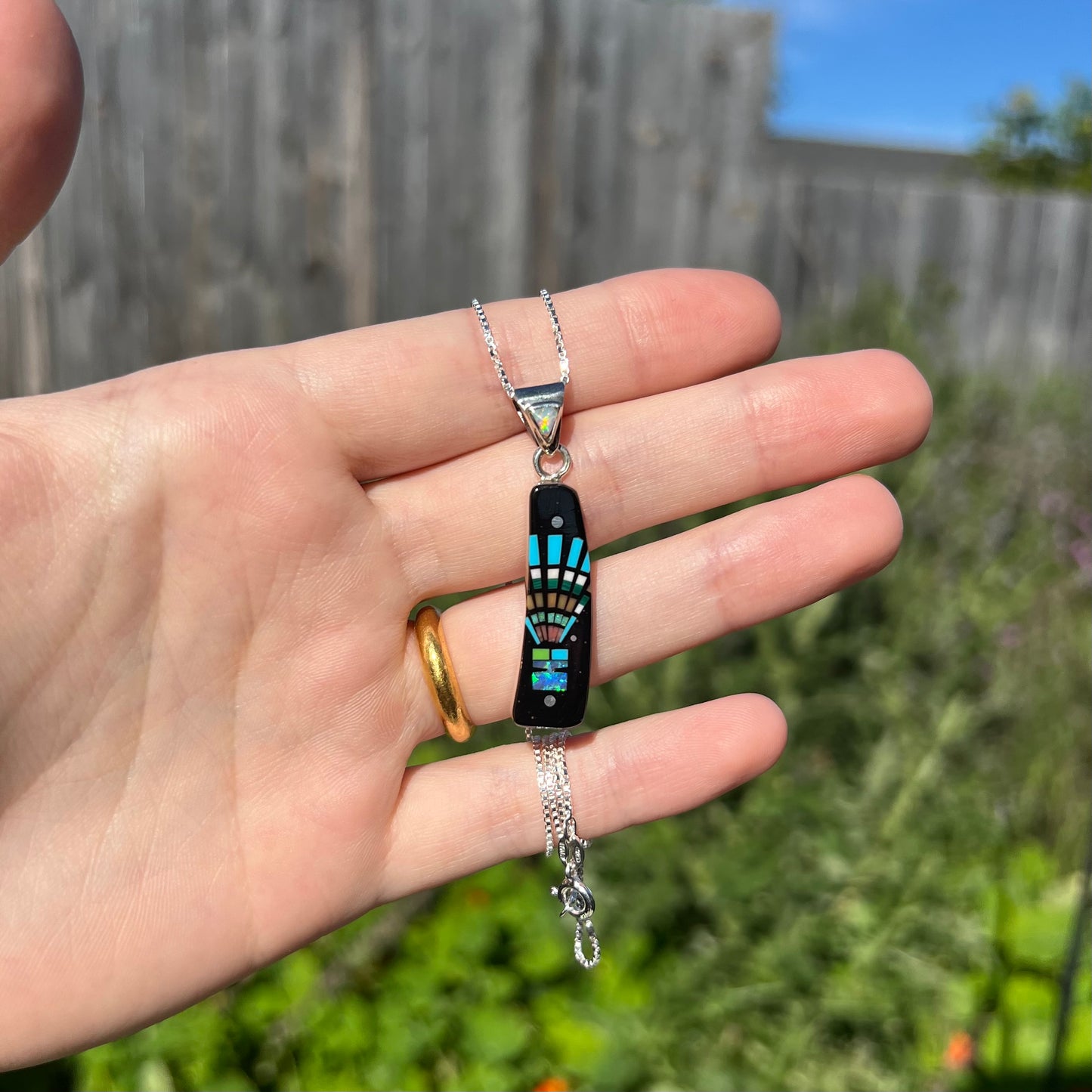 Hand holding Southwest-style multi-stone inlay pendant in sunlight against blurred outdoor background.