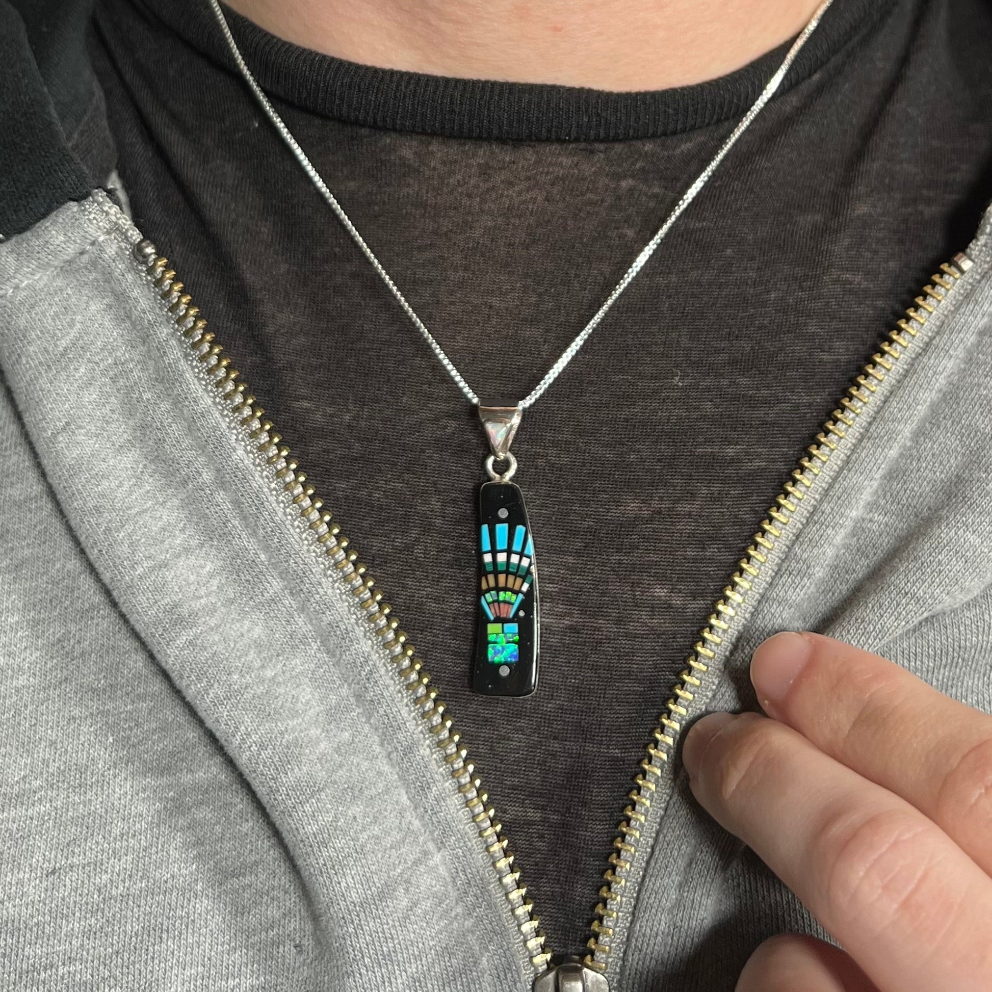 Man wearing Southwest-style sterling silver inlay necklace with kachina motif around neck under unzipped hoodie on dark shirt.