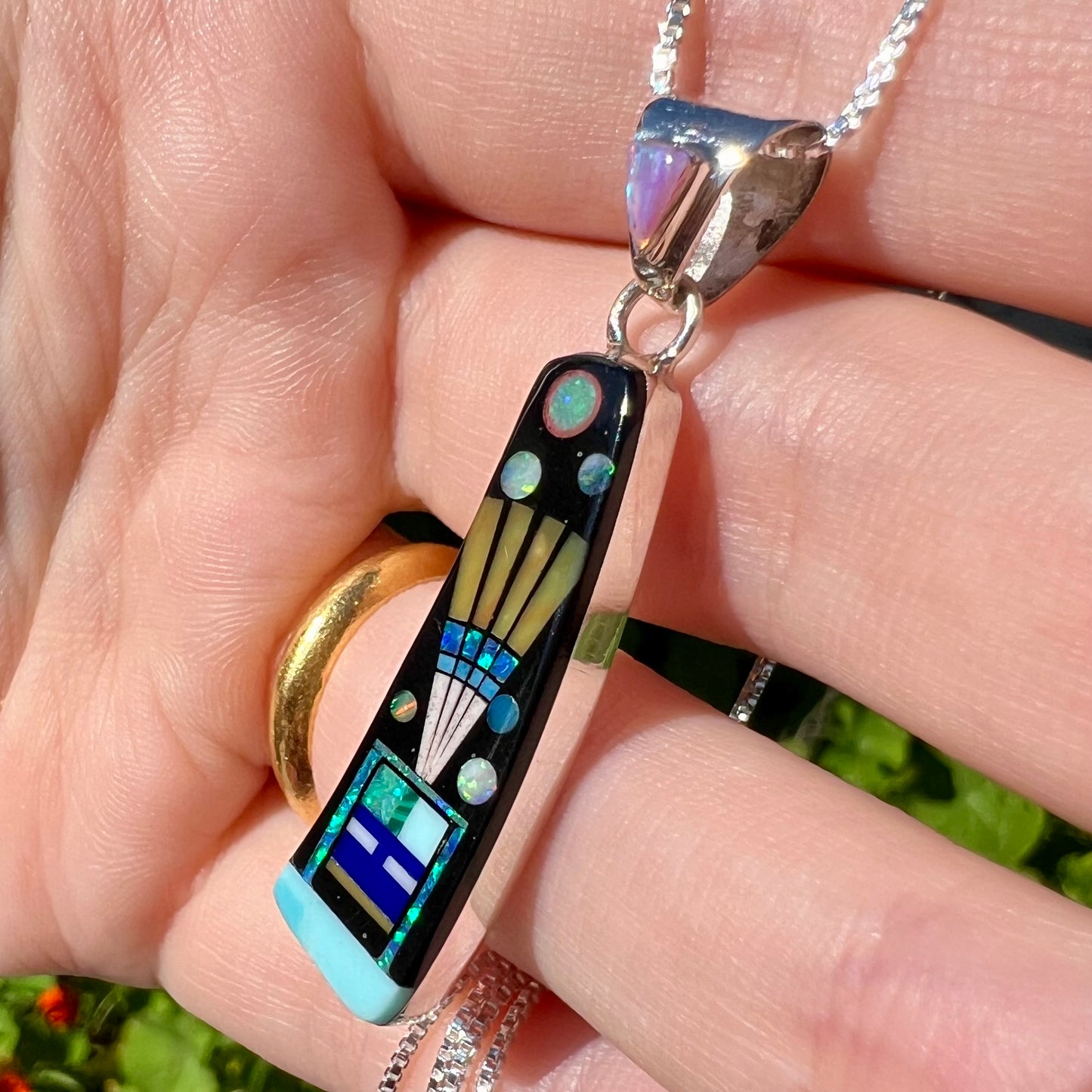 Men's Southwest-style stone inlay pendant with kachina in headdress design, held in a hand and shown in sunlight close-up from angle.