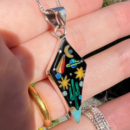 Southwest-style opal and onyx inlay pendant shown from angle in hand.  A blue opal comet has a red tail in natural light.