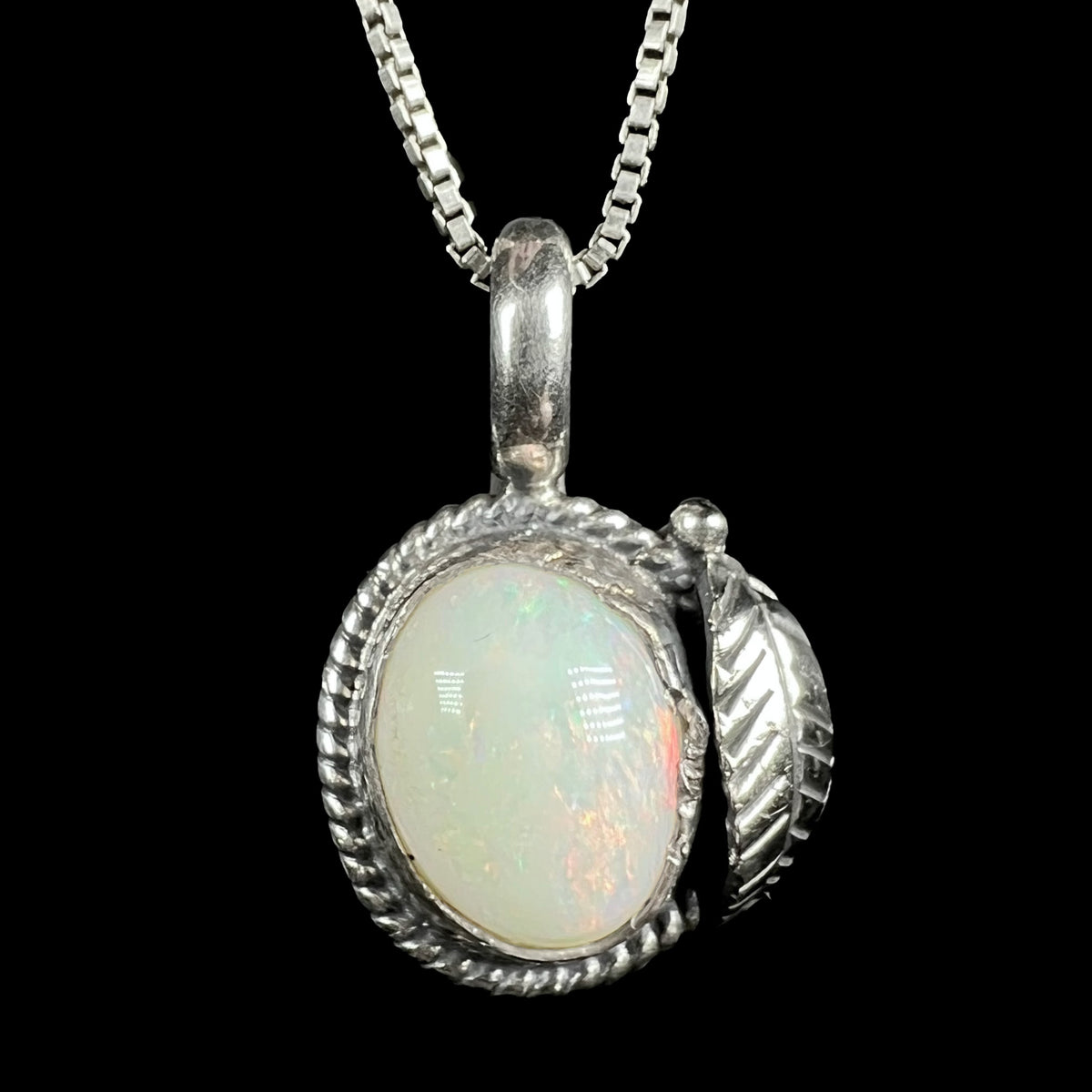 A sterling silver pendant necklace featuring an oval white opal gemstone with iridescent color flashes, set in a Southwest inspired textured rope-style bezel with a feather-shaped charm on the side, suspended from a silver box chain against a solid black background.