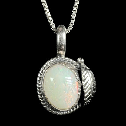 A sterling silver pendant necklace featuring an oval white opal gemstone with iridescent color flashes, set in a Southwest inspired textured rope-style bezel with a feather-shaped charm on the side, suspended from a silver box chain against a solid black background.