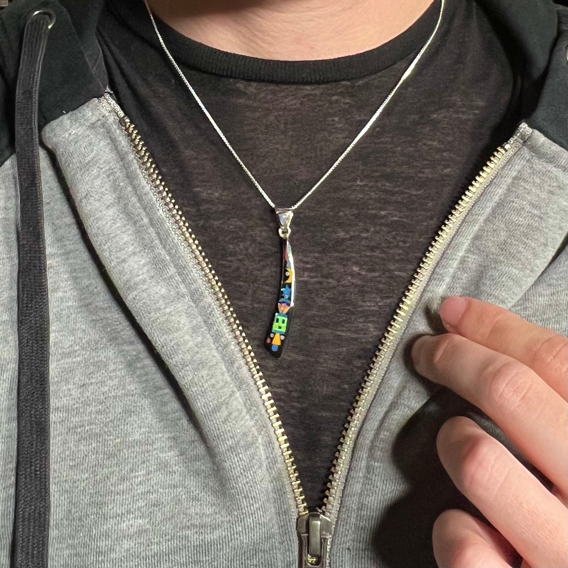 Person wearing elongated Southwest-style sterling silver inlay necklace around neck under unzipped hoodie against dark shirt.