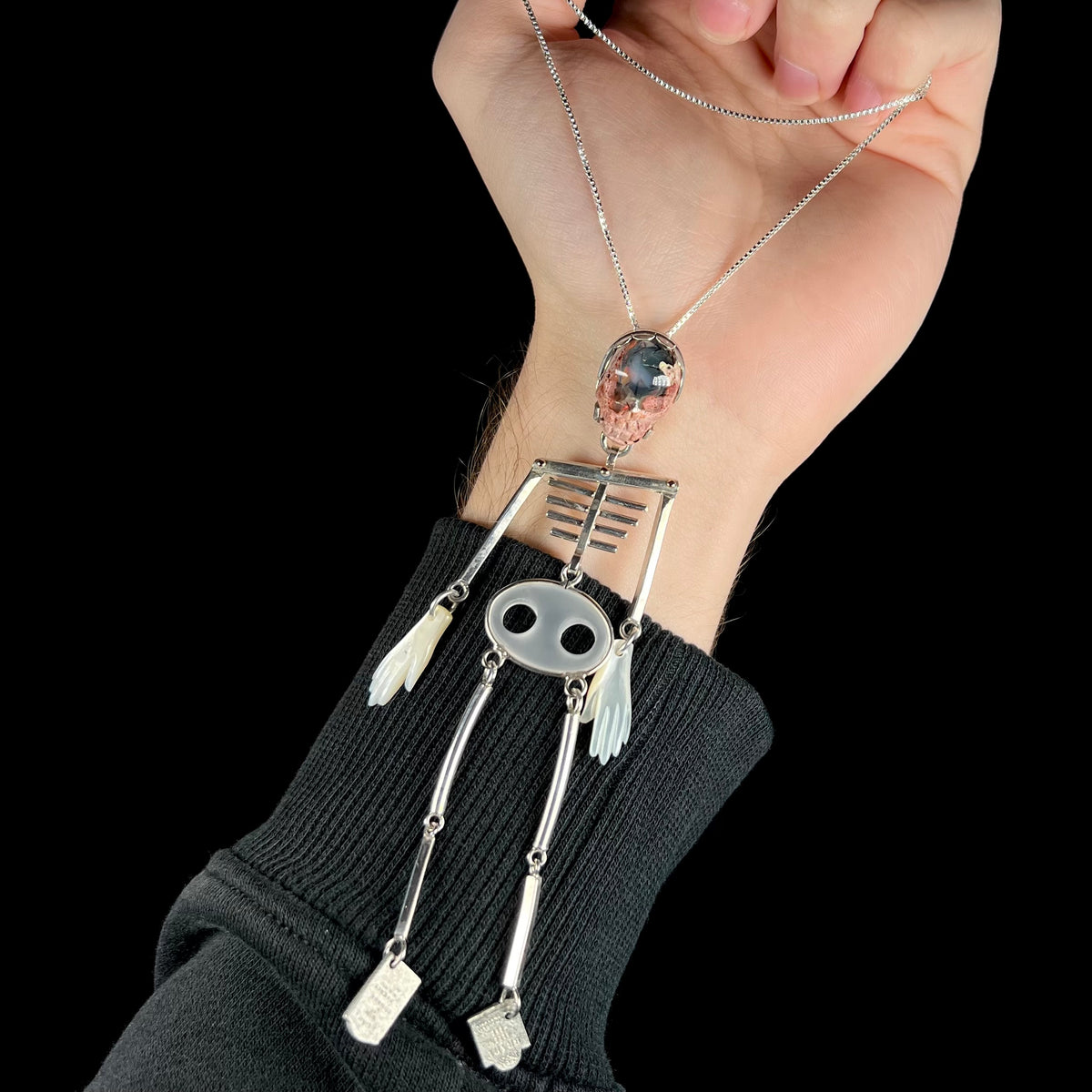 A hand wearing a black knit sweater holds up a silver skeleton pendant necklace from with a colorful cantera opal skull and mother of pearl hands from chain against a solid black background.