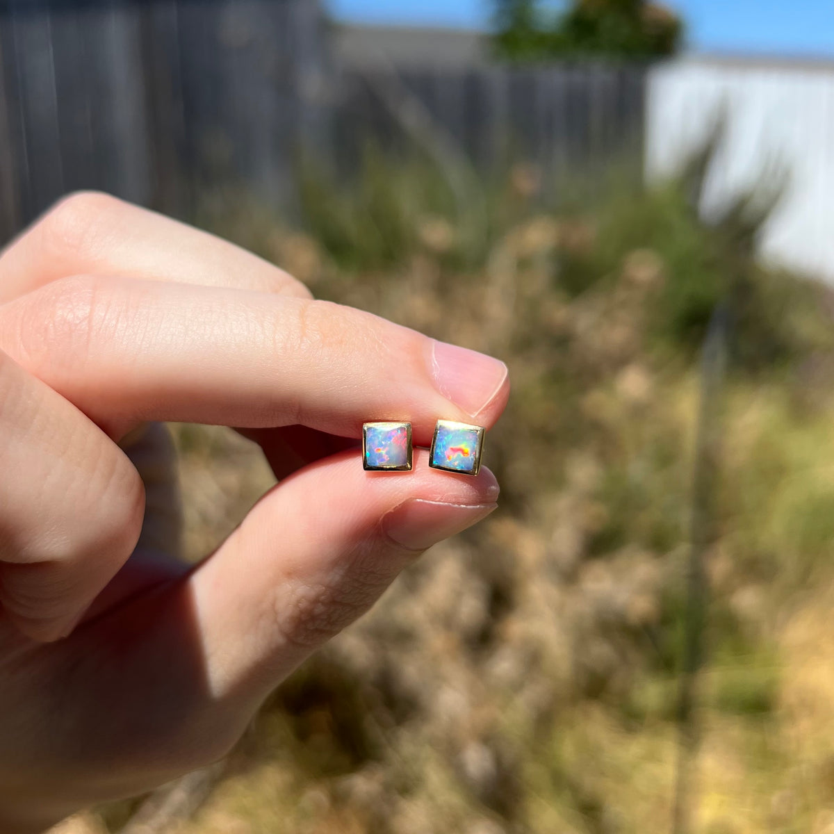 Square shaped opal stud earrings with red flashes held between fingers in natural light