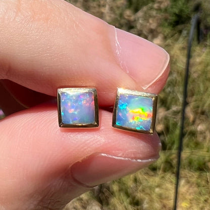 Close-up of square cut opal stud earrings in 14k gold, shown in sunlight held between two fingers