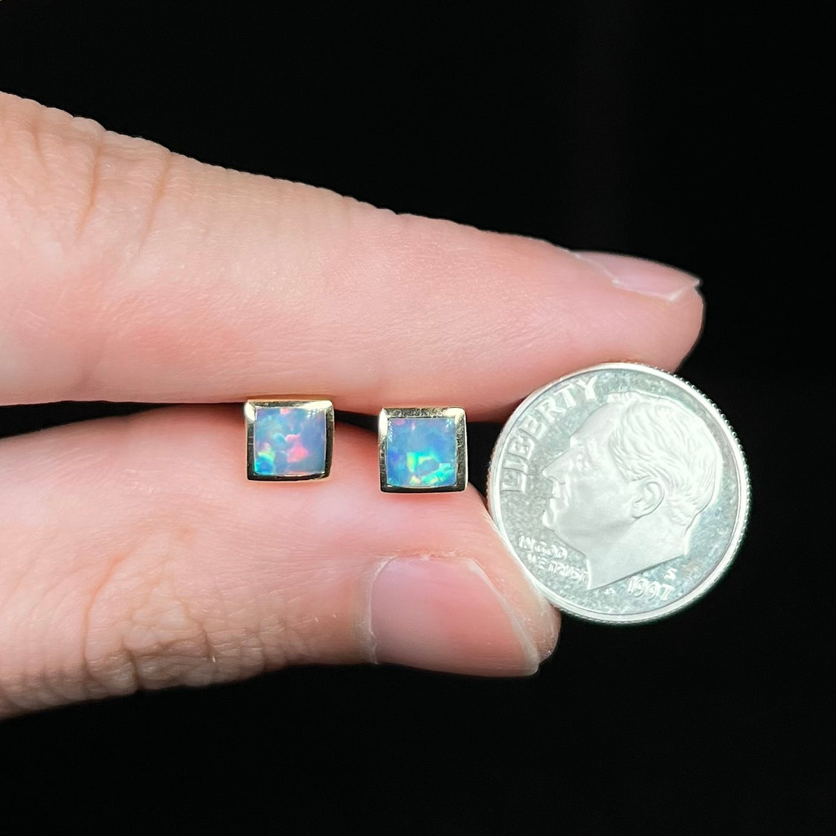 Fingers holding pair of square shaped opal stud earrings next to US dime coin for scale