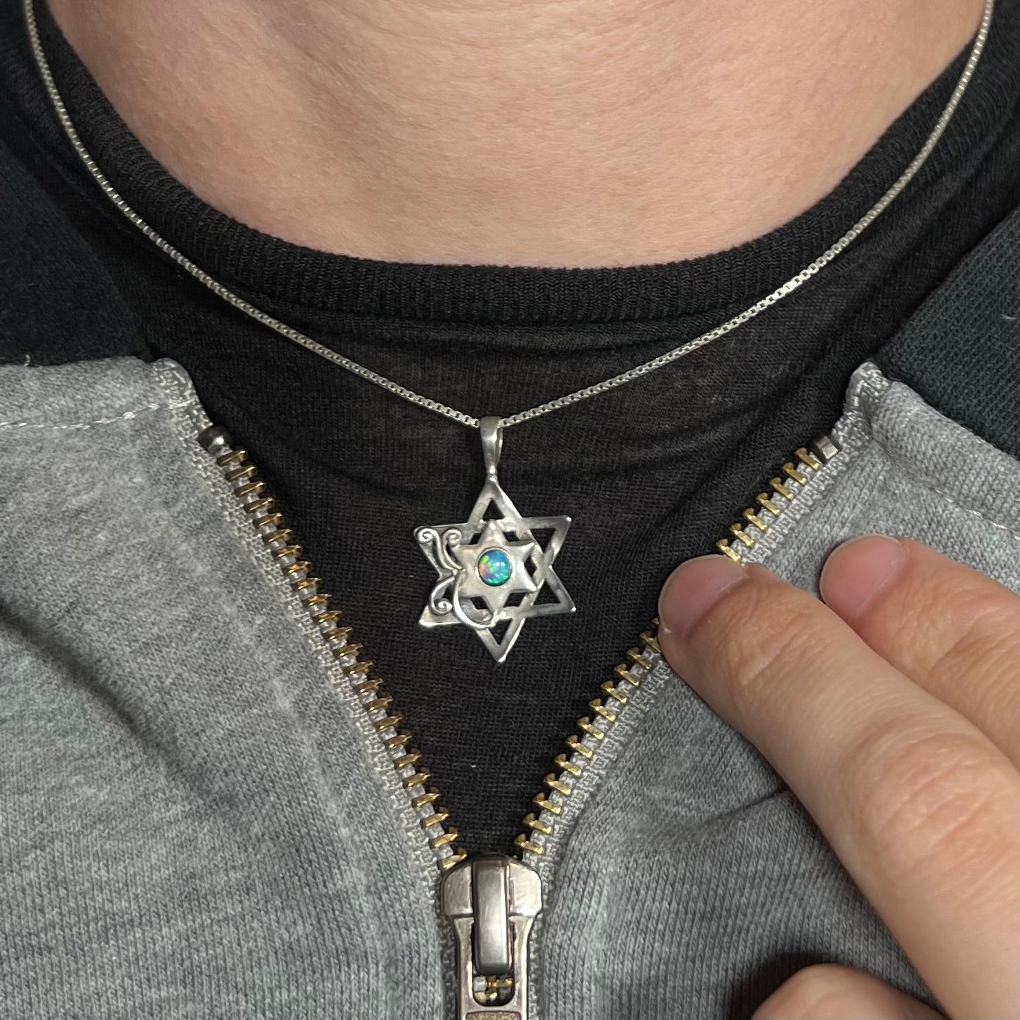 A sterling silver Star of David opal necklace worn against a dark shirt with a partially unzipped jacket.