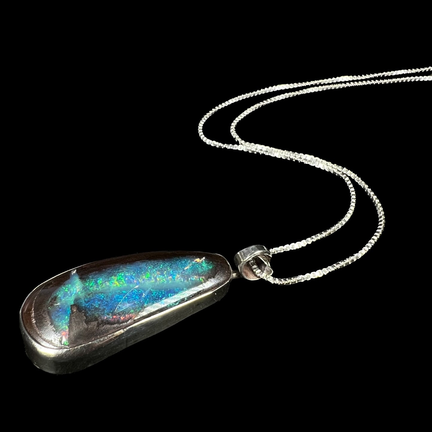 A sterling silver boulder opal pendant with glittering blue and green color play, shown from an angle laying on black.