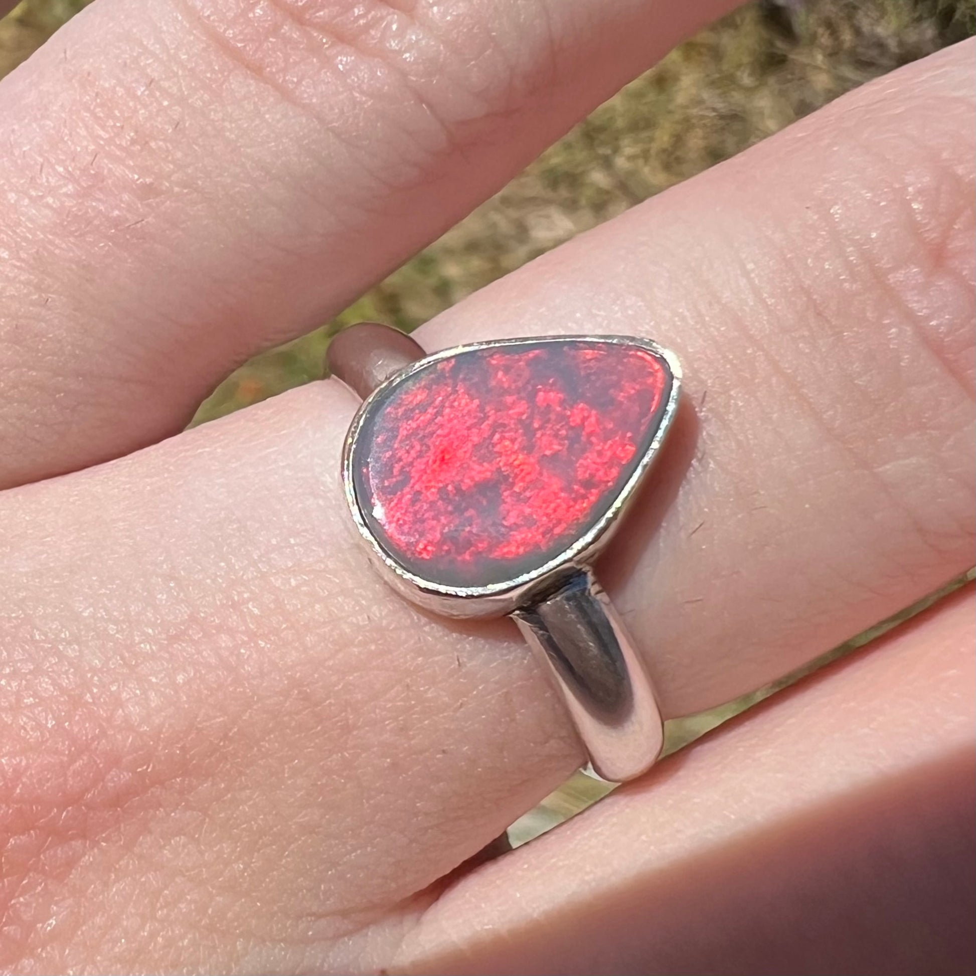 Close-up of a sterling silver pear shaped Australian opal women's ring with a broad neon-red flash of color in the sunlight, worn on a hand.