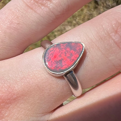 Close-up of a sterling silver pear shaped Australian opal women's ring with a broad neon-red flash of color in the sunlight, worn on a hand.