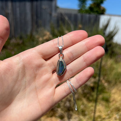 Hand holding a sterling silver black opal men's pendant in the sunlight.  Three green flashes are seen along the opal's right edge.