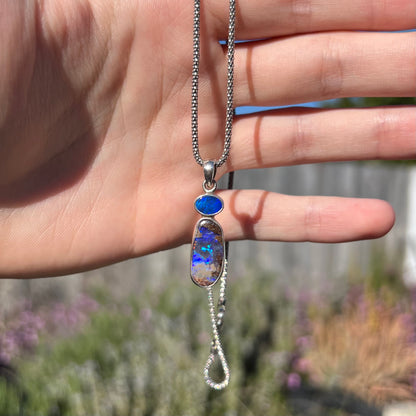 A sterling silver boulder opal necklace dangling from a hand in natural sunlight, revealing a greenish-blue opal flash.