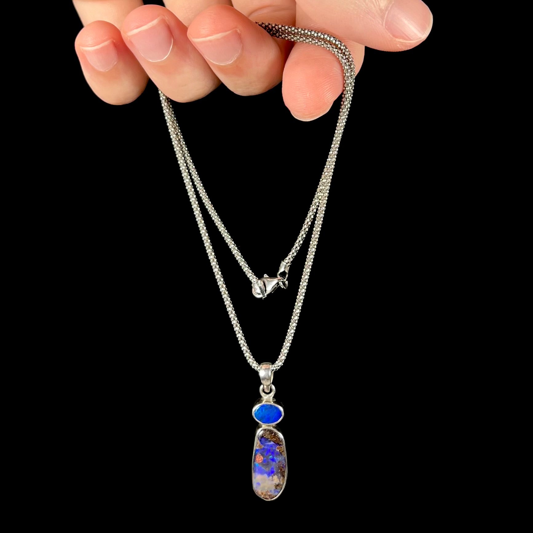 A sterling silver boulder opal necklace dangling from a hand, displaying a vibrant red flash in the opal’s deep blue body tone against a black background.