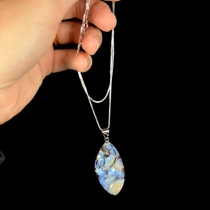 A large, blue Australian boulder opal necklace in sterling silver, dangled by its chain from a hand.