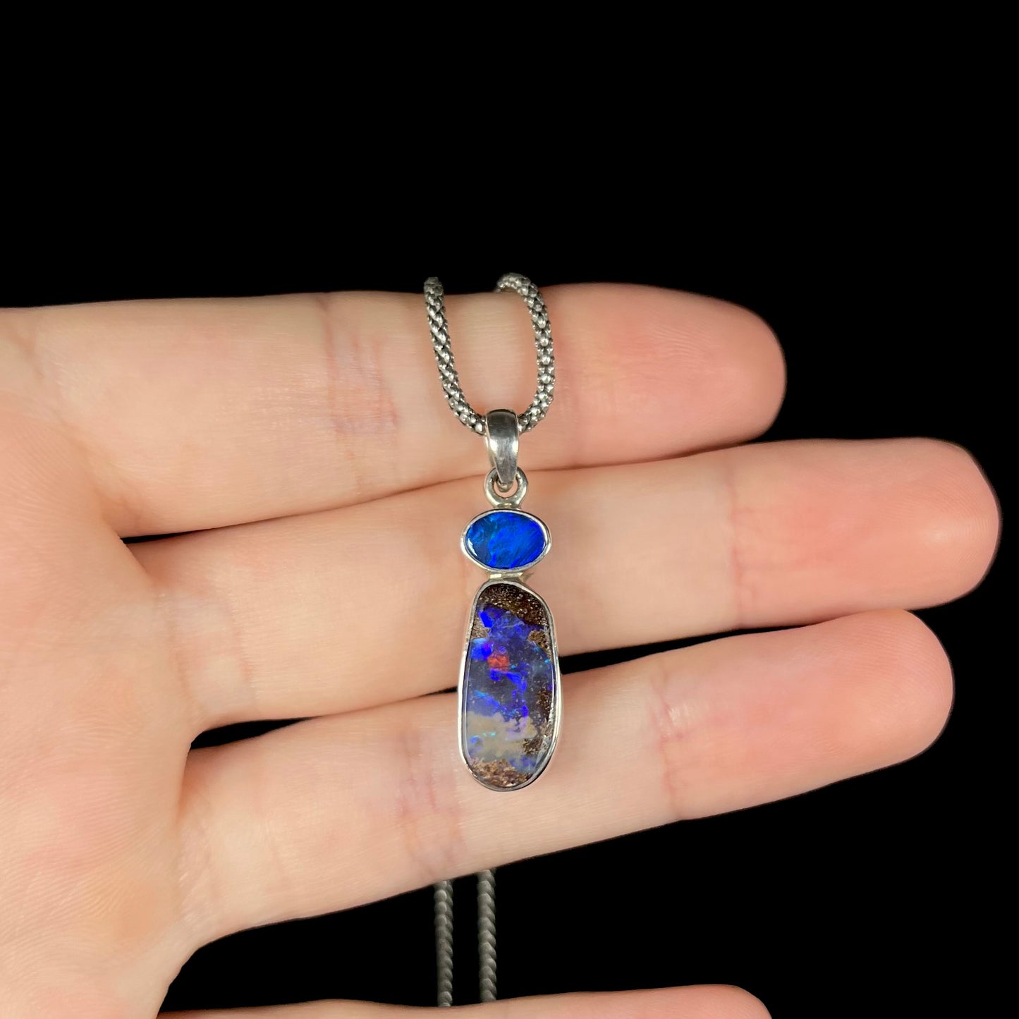 A sterling silver boulder opal necklace resting on fingers against a black background, featuring a deep blue opal with a strong red flash.