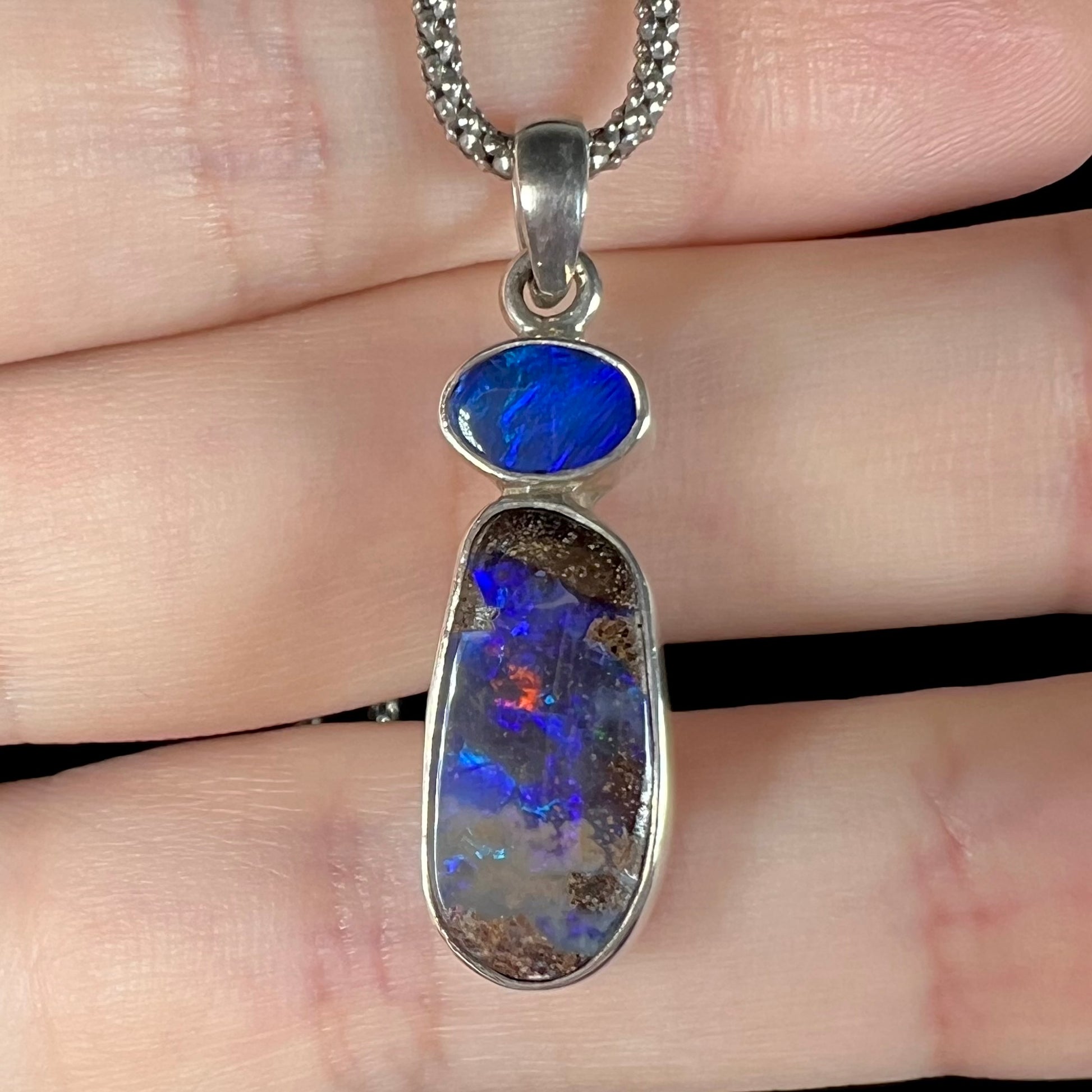 A macro close-up of a sterling silver boulder opal necklace resting on fingers, highlighting the opal’s red flash against its deep blue body tone.