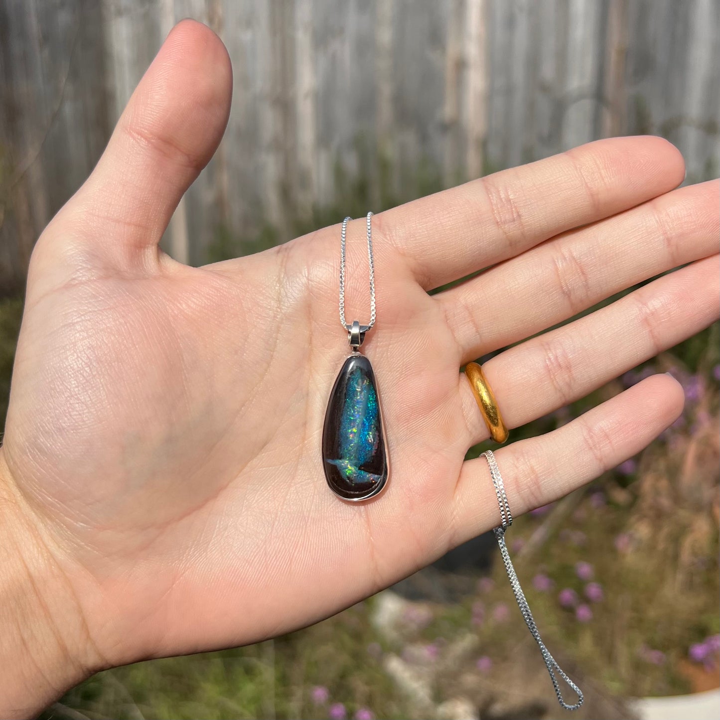A sterling silver boulder opal necklace with glittering blue color play, shown in a hand in sunlight.