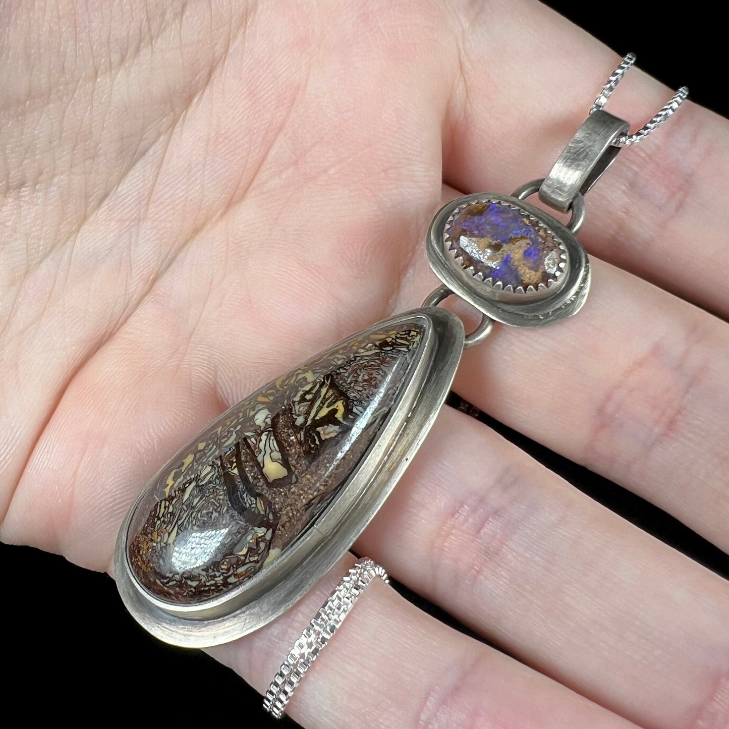 Unisex sterling silver boulder opal necklace shown from an angle close-up held in a hand under cool studio lights.