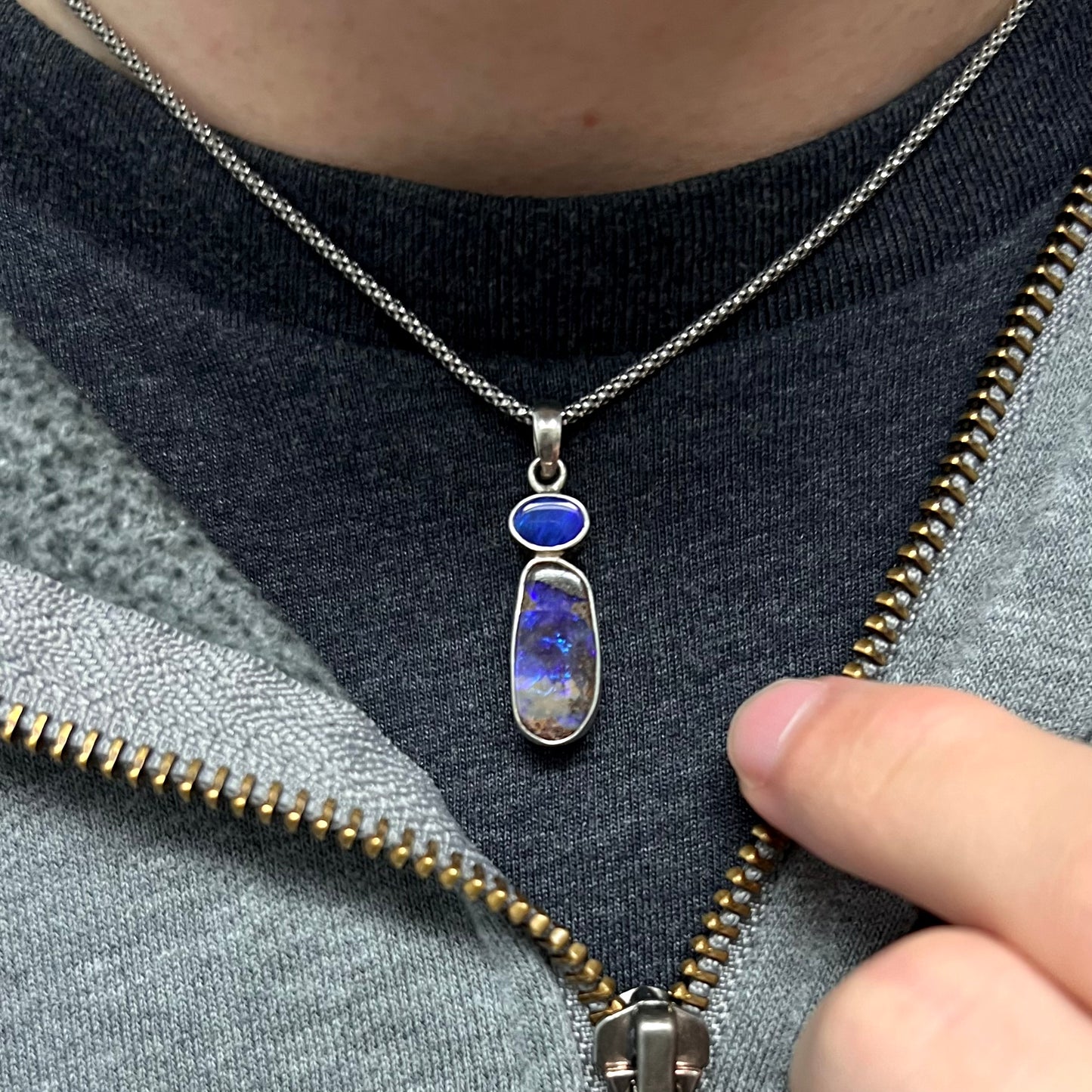 A sterling silver boulder opal necklace worn around the neck on a black t-shirt, with a partially zipped sweatshirt, revealing only a blue opal flash.