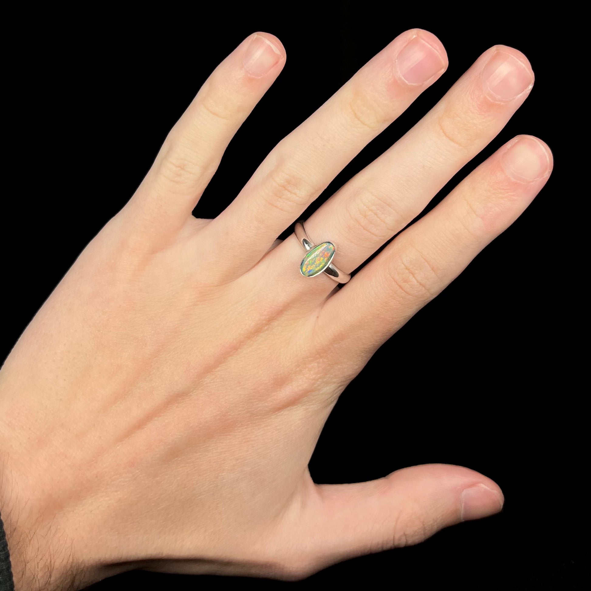 Hand wearing a women's sterling silver Australian dark opal solitaire ring under cool LED studio light.  The opal has orange and red play of color.
