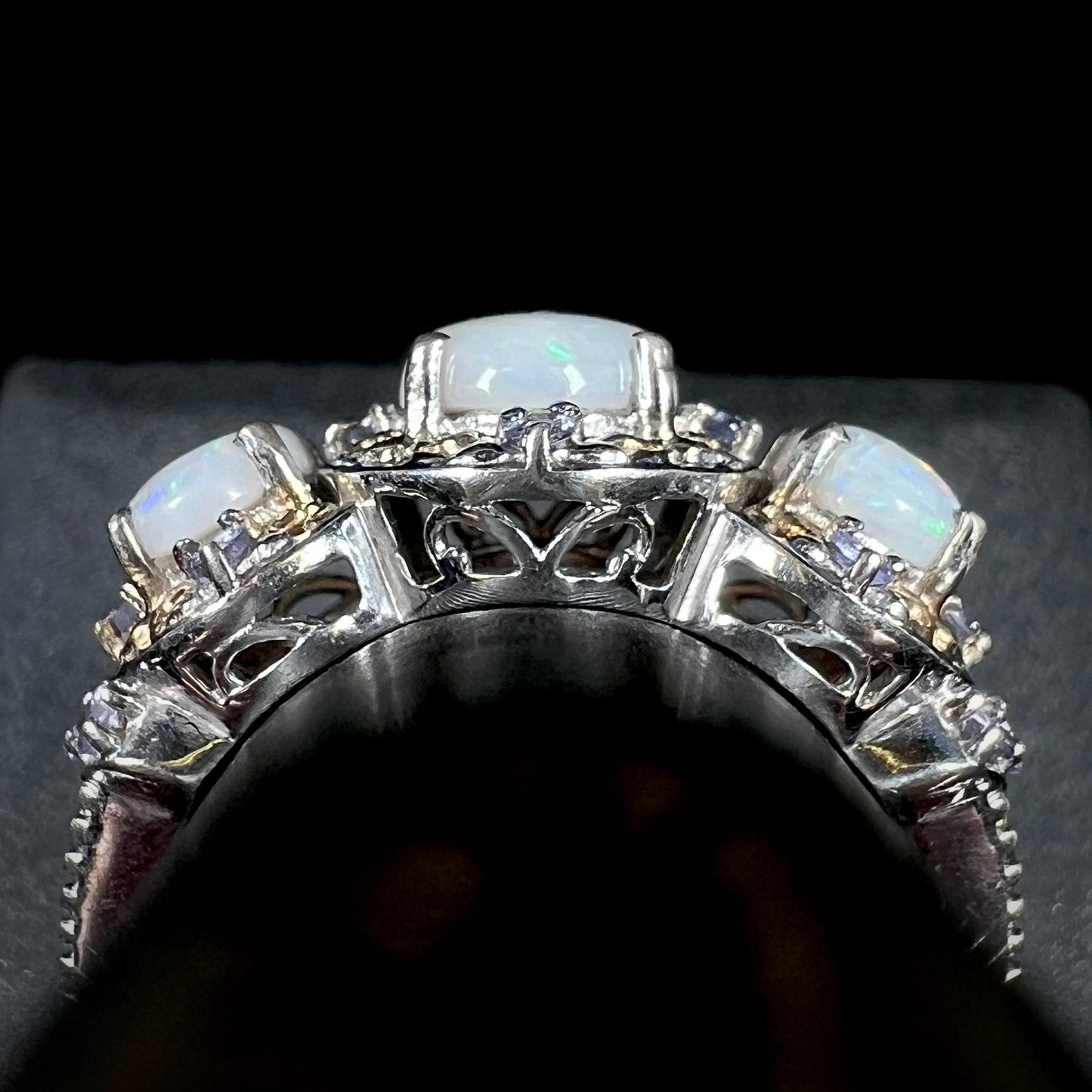Side gallery profile view of filigree details on the basket of a sterling silver three-stone opal and tanzanite ring, shown on a ring display.
