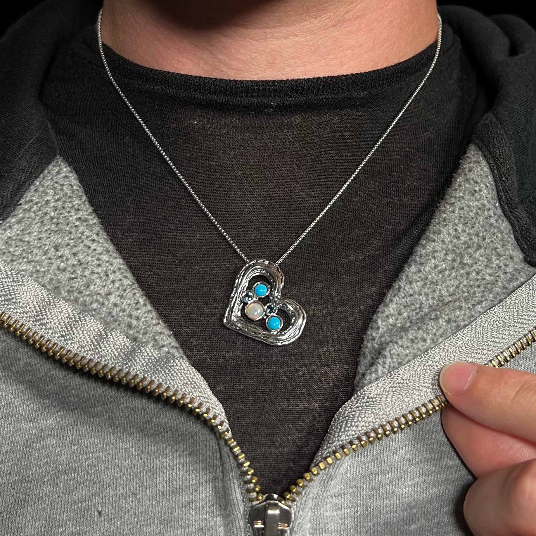 A sterling silver heart necklace set with opal, turquoise, and blue topaz, worn around a neck on a dark shirt.