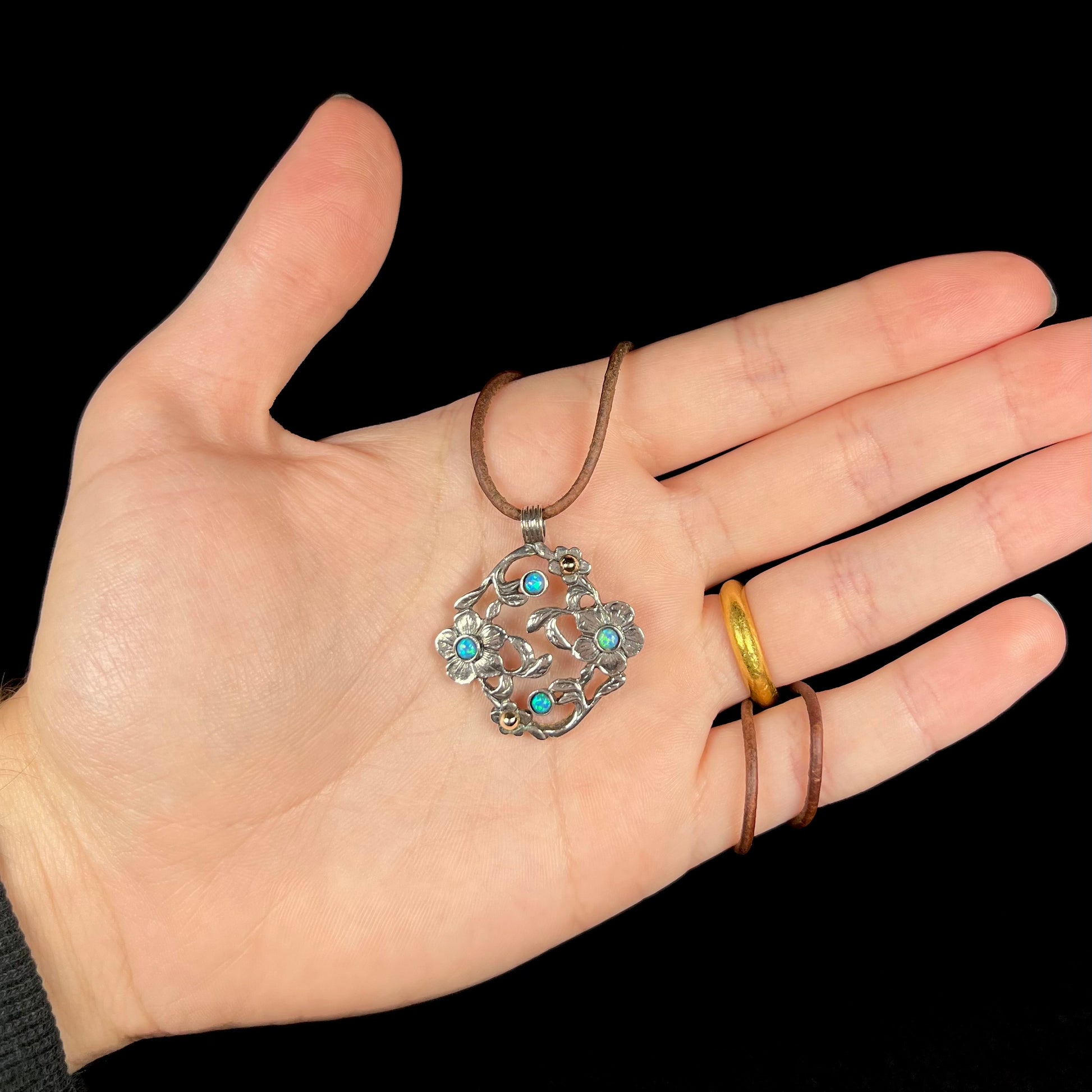 A two-tone sterling silver and 14k gold opal dogwood flower necklace on a brown cord in a hand against black background.