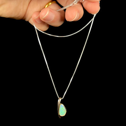 A green Australian crystal opal necklace in sterling silver, shown dangled by its chain from a hand against a black background.