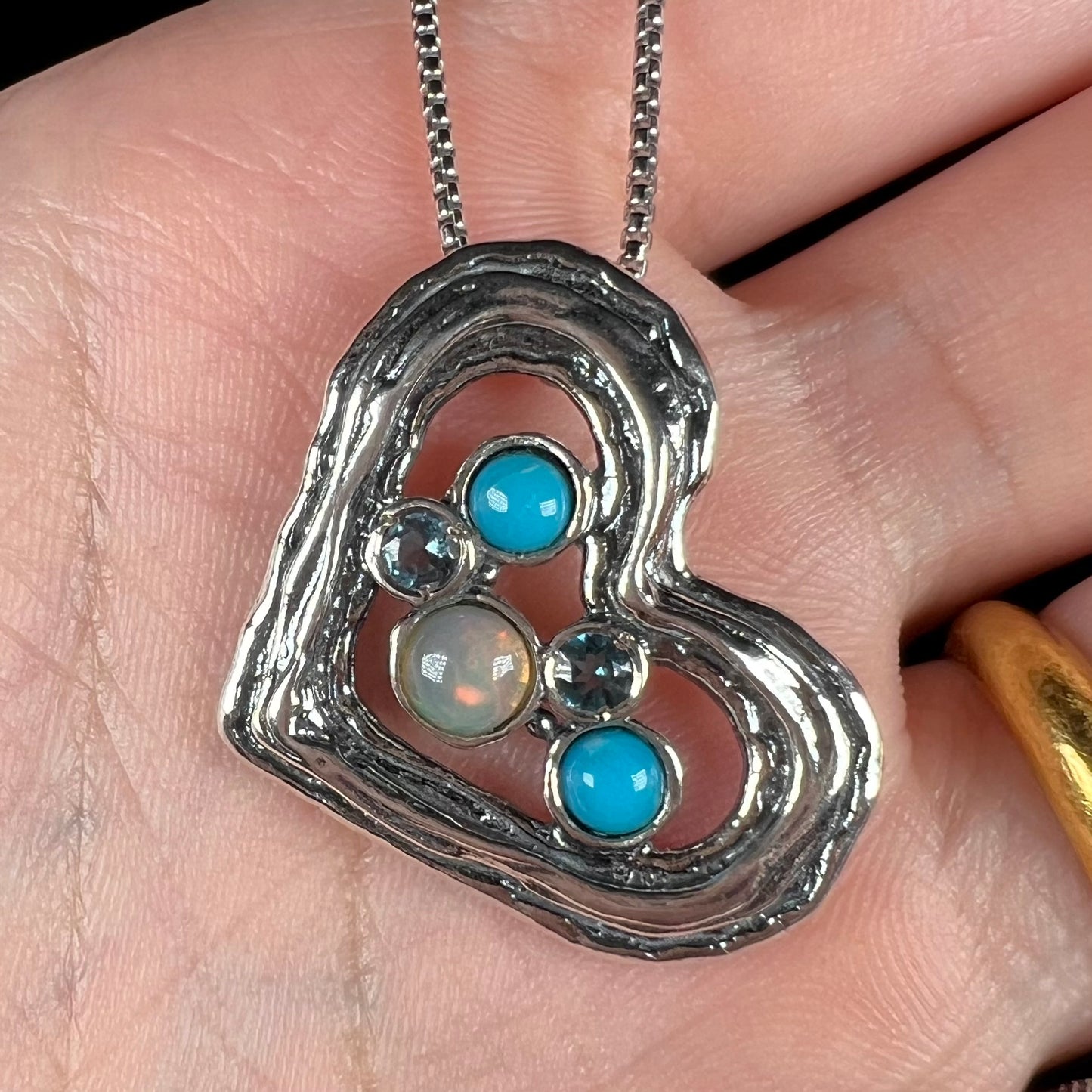 Close-up of a sterling silver heart necklace set with opal, turquoise, and blue topaz in a hand.