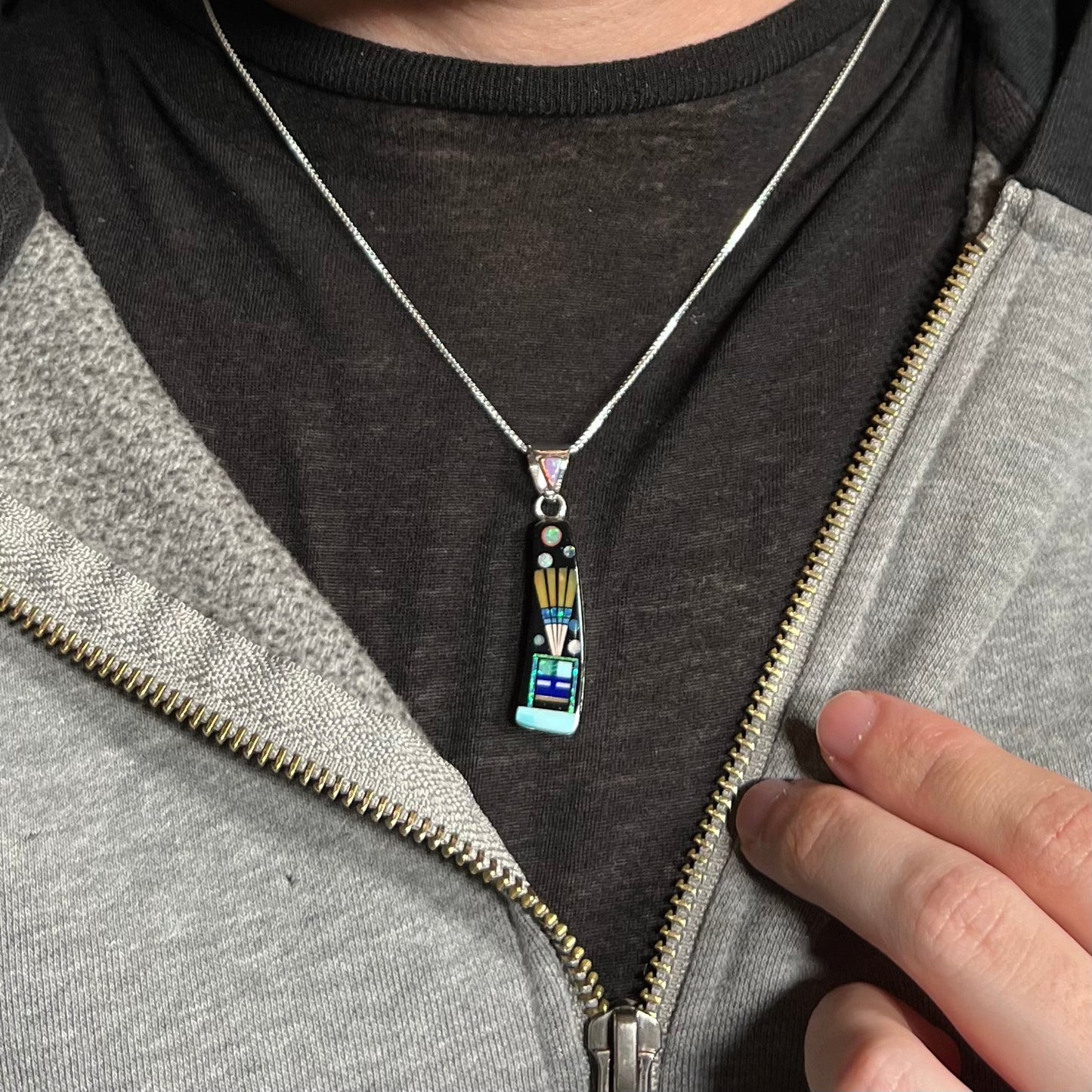 Man wearing sterling silver inlay necklace with kachina motif around neck under unzipped hoodie on dark shirt.