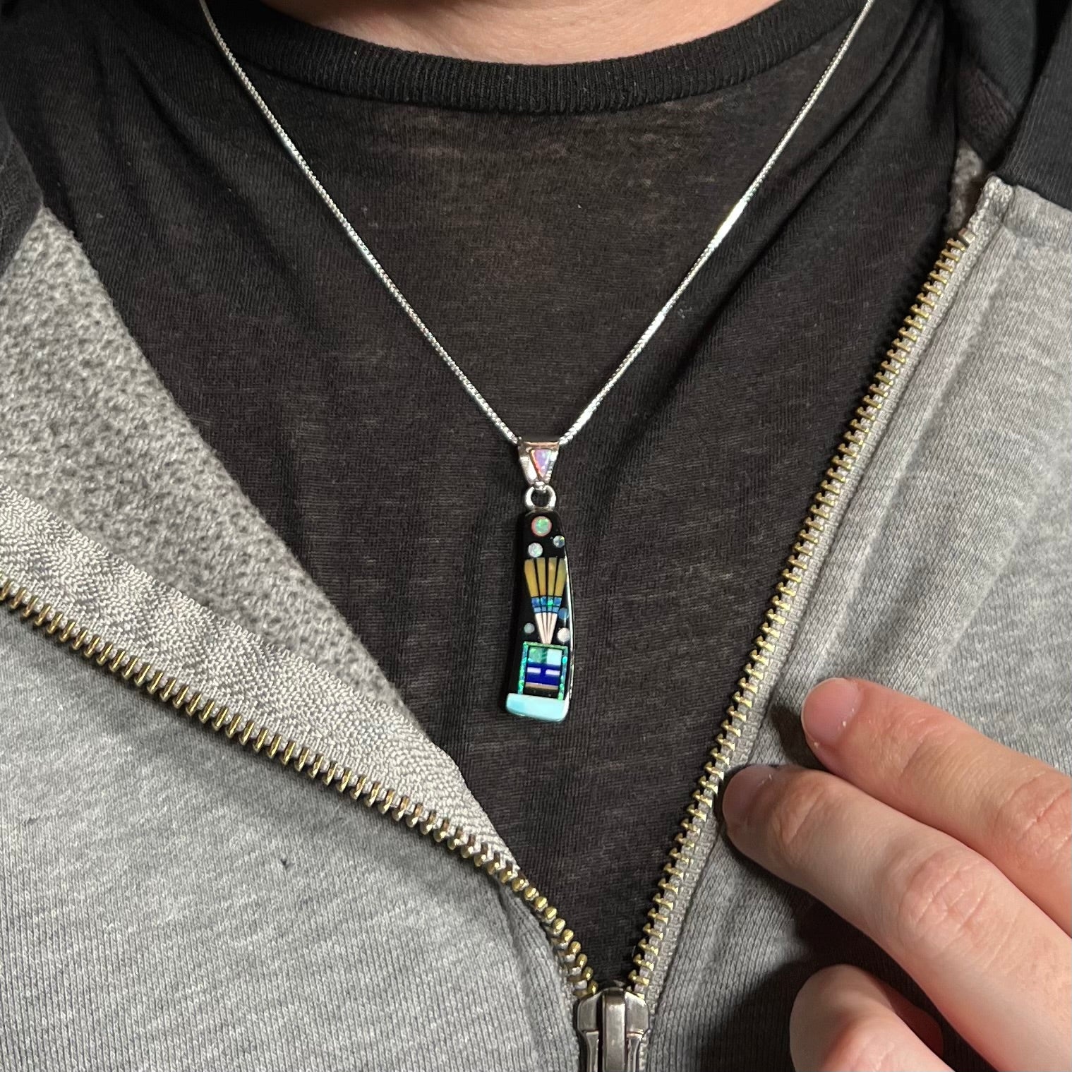 Man wearing sterling silver inlay necklace with kachina motif around neck under unzipped hoodie on dark shirt.
