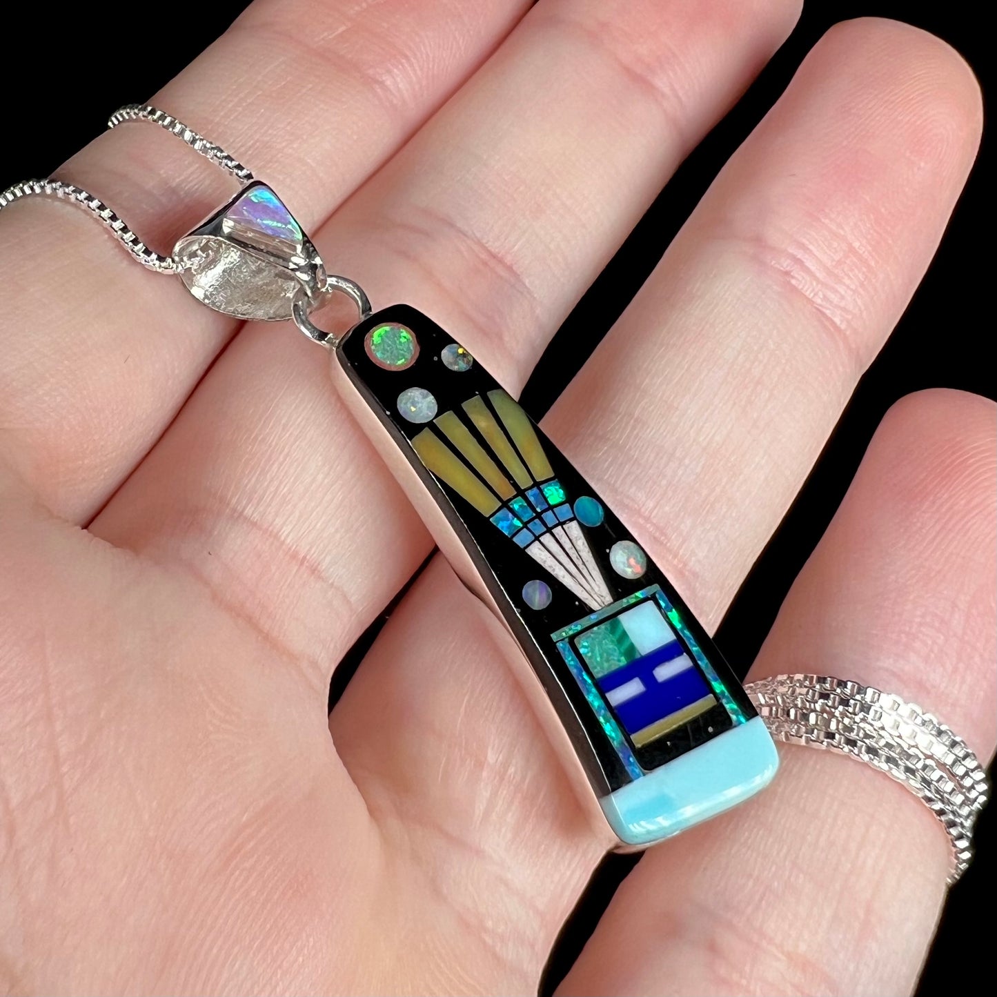 Angled close-up of sterling silver opal and onyx inlay pendant with kachina in headdress design, held in hand under cool studio light.
