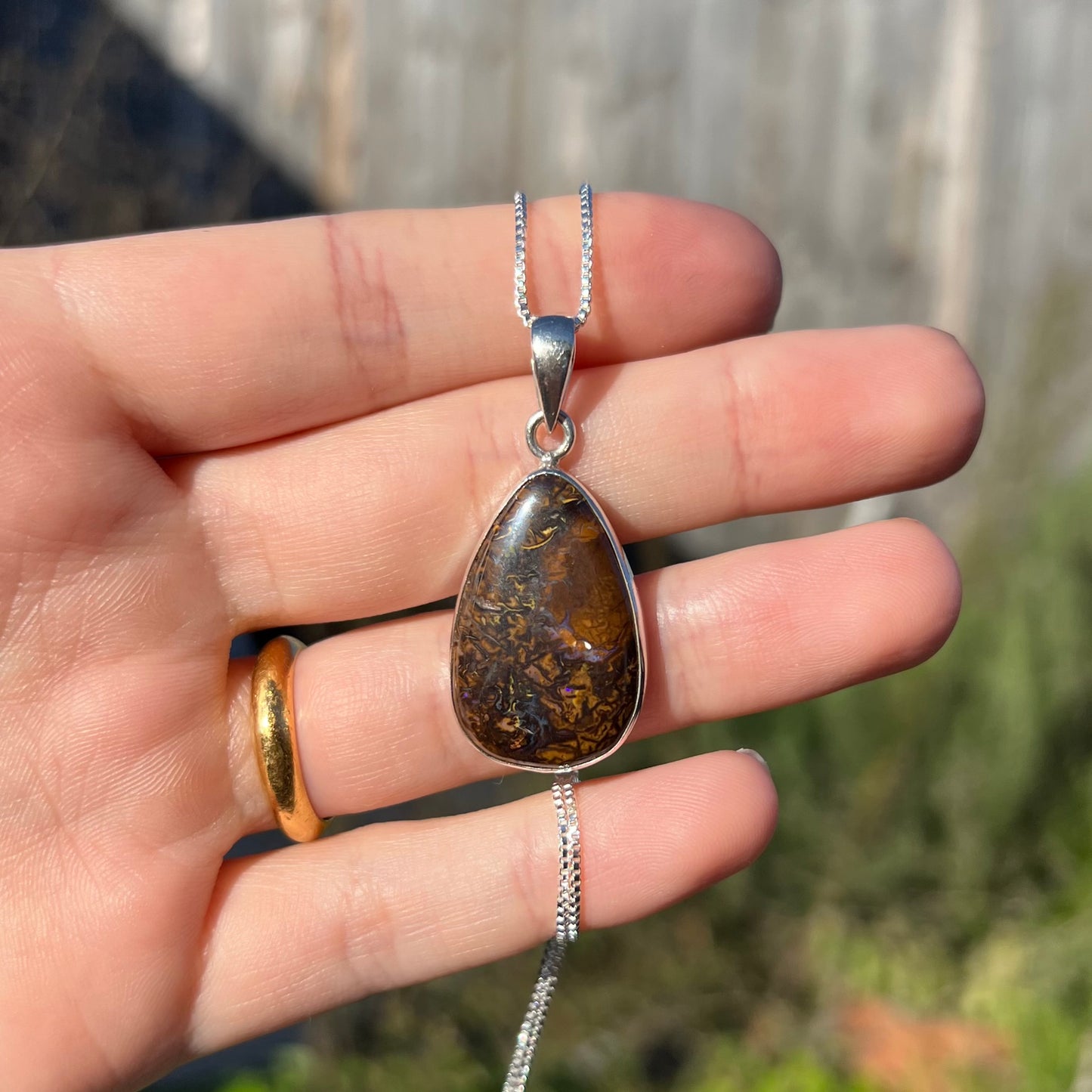 A pear shaped Koroit boulder opal pendant in sterling silver shown in a hand in the sunlight.