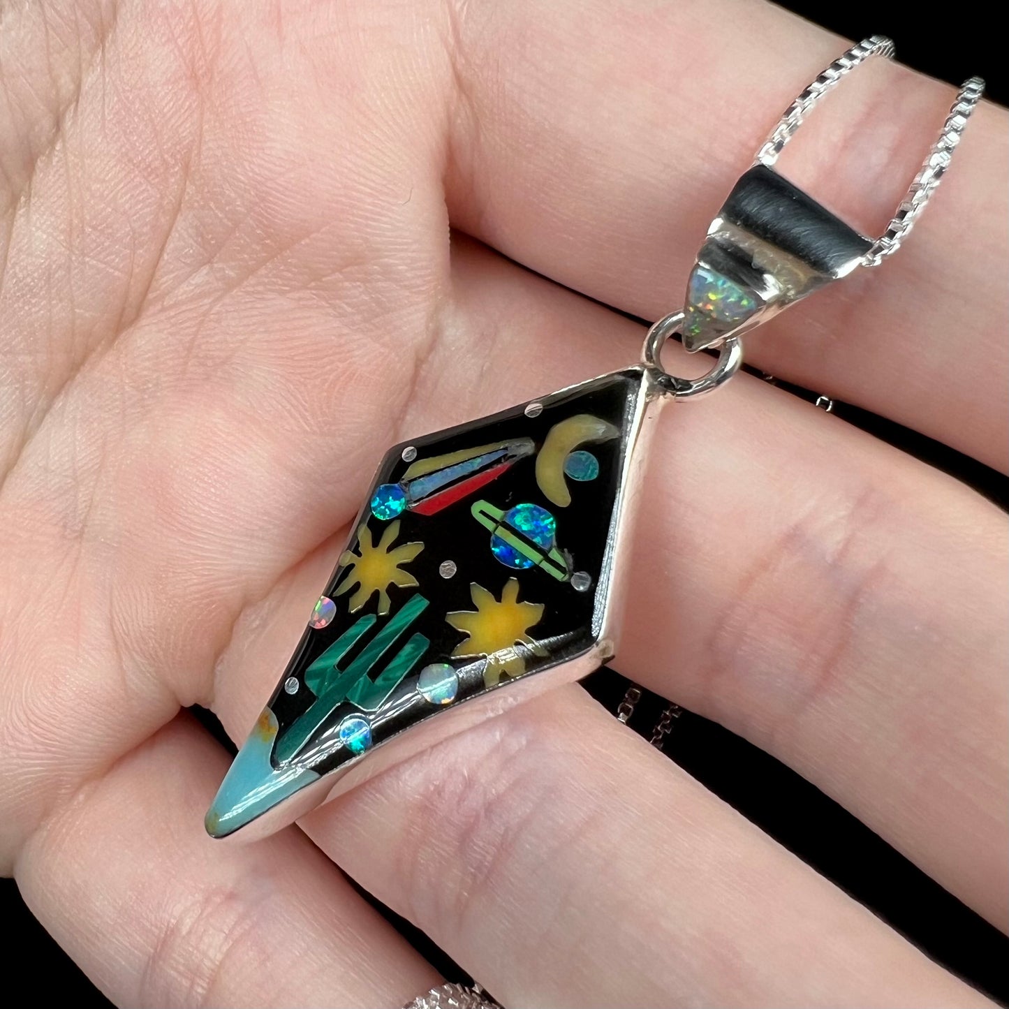 Angled close-up of sterling silver lab-created opal and onyx inlay pendant with outer-space motif, held in a hand.