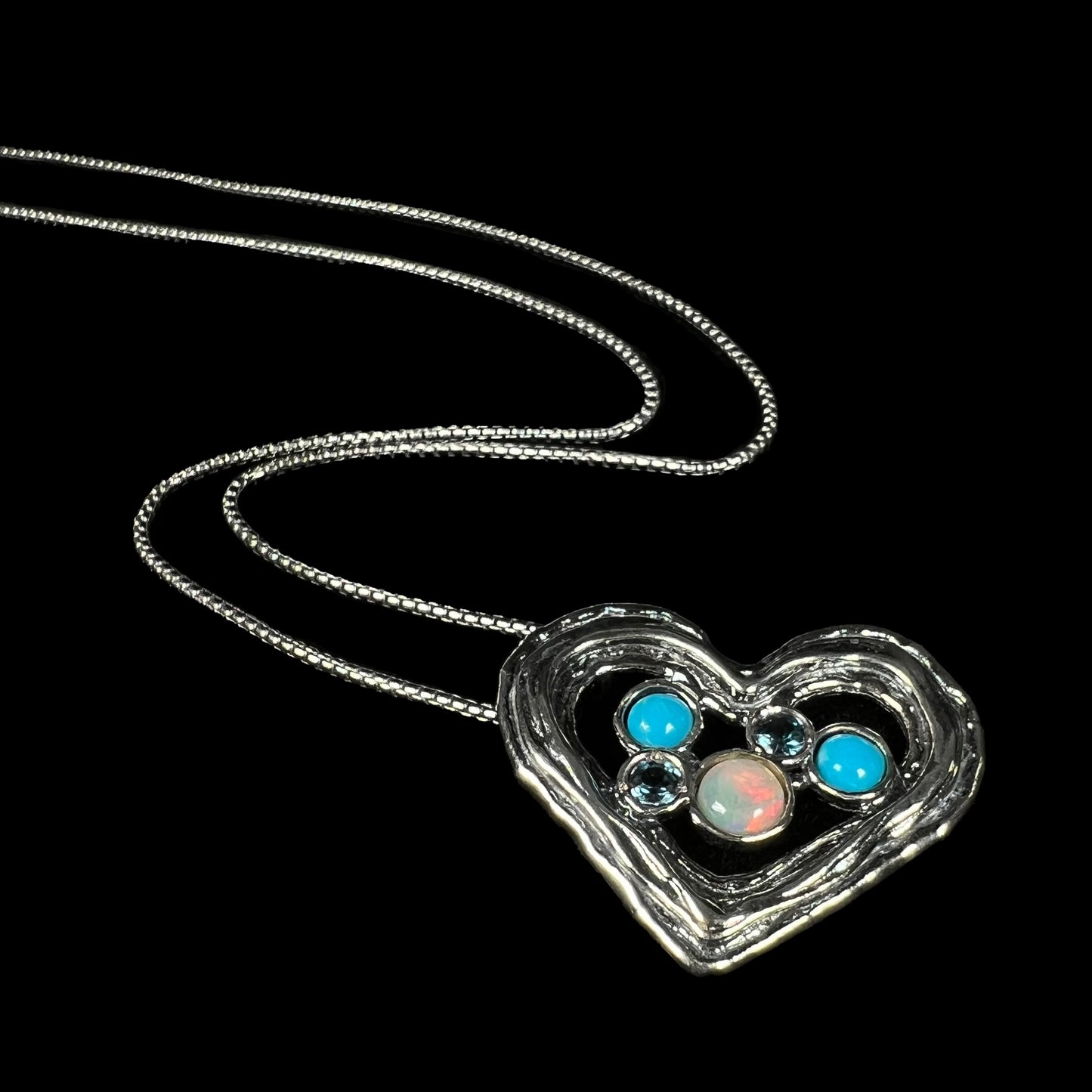 A silver heart necklace with Ethiopian opal, turquoise, and blue topaz, shown from angle on black backdrop.