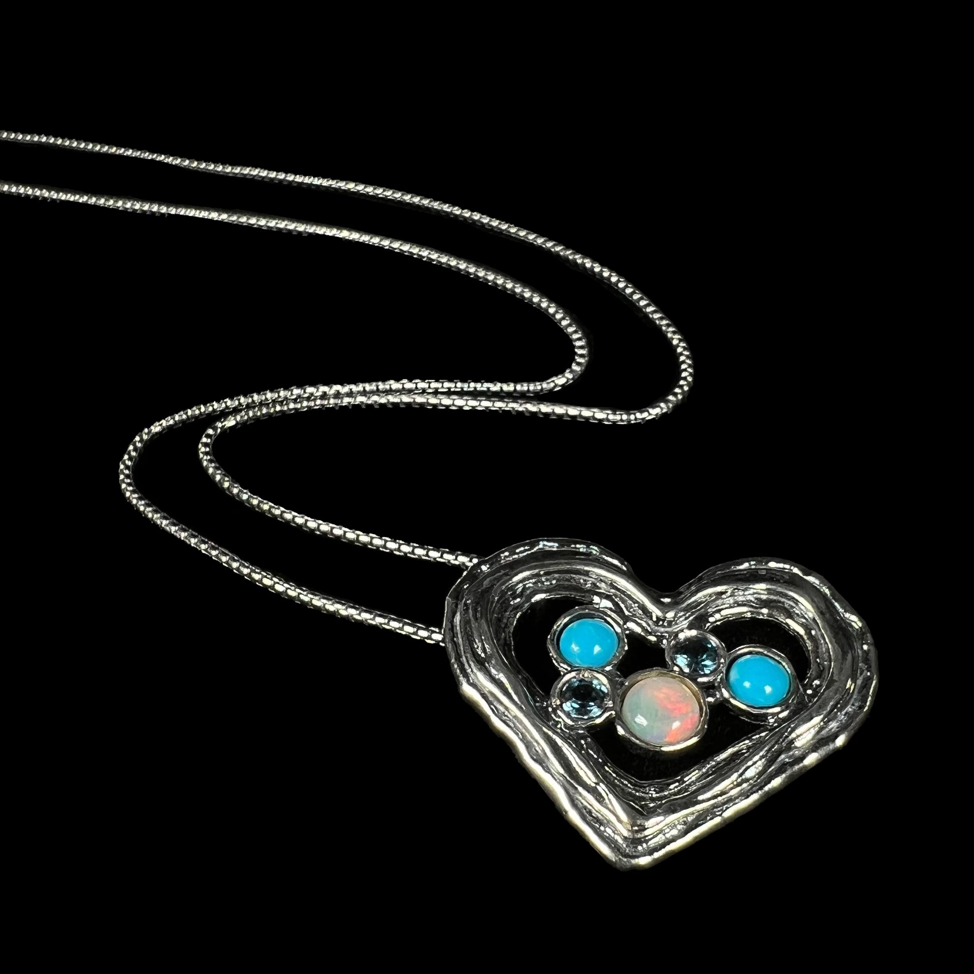 A silver heart necklace with Ethiopian opal, turquoise, and blue topaz, shown from angle on black backdrop.