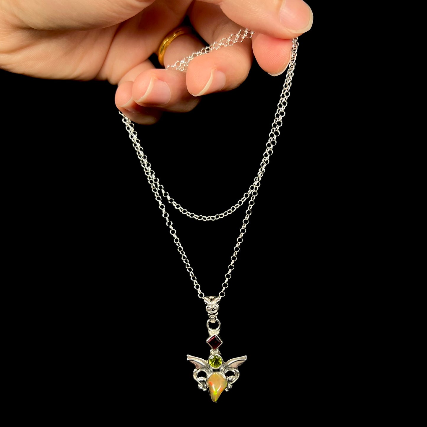 A sterling silver angel necklace set with an fire opal, peridot, and garnet, dangled from a hand by its chain.