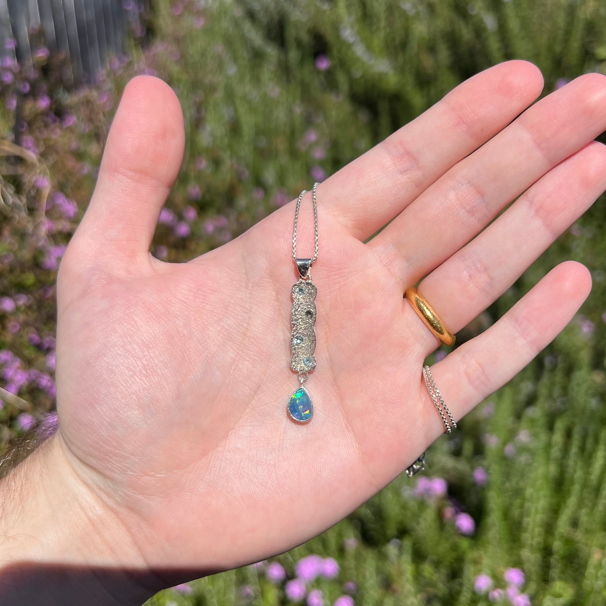 Hand holding a sterling silver Australian opal drop necklace with topaz accents in natural sunlight against blurred green background.