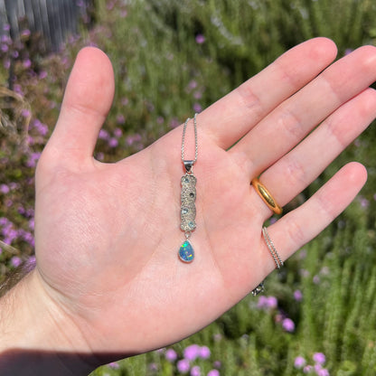 Hand holding a sterling silver Australian opal drop necklace with topaz accents in natural sunlight against blurred green background.