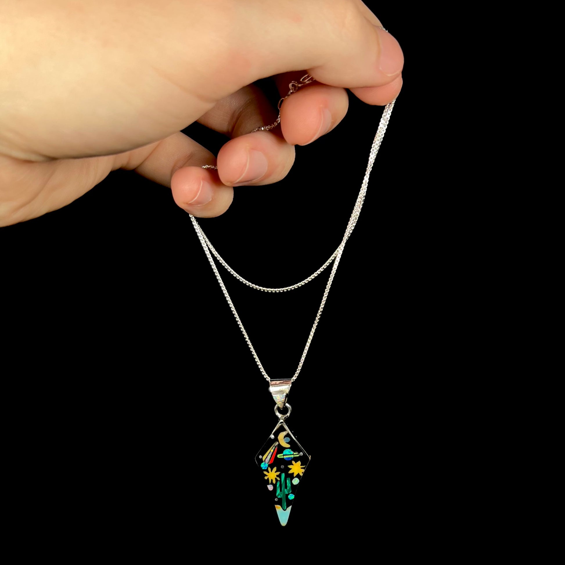 Sterling silver opal and onyx inlay necklace with Southwest space-inspired motif dangled from chain by hand in front of black background.